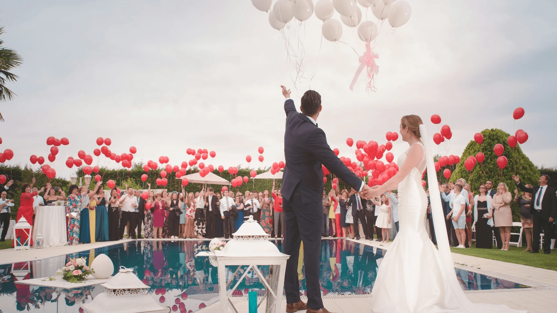 A bride and groom stand hand-in-hand by a pool, releasing white balloons into a cloudy sky as wedding guests holding red balloons watch and celebrate around them at an outdoor villa reception.