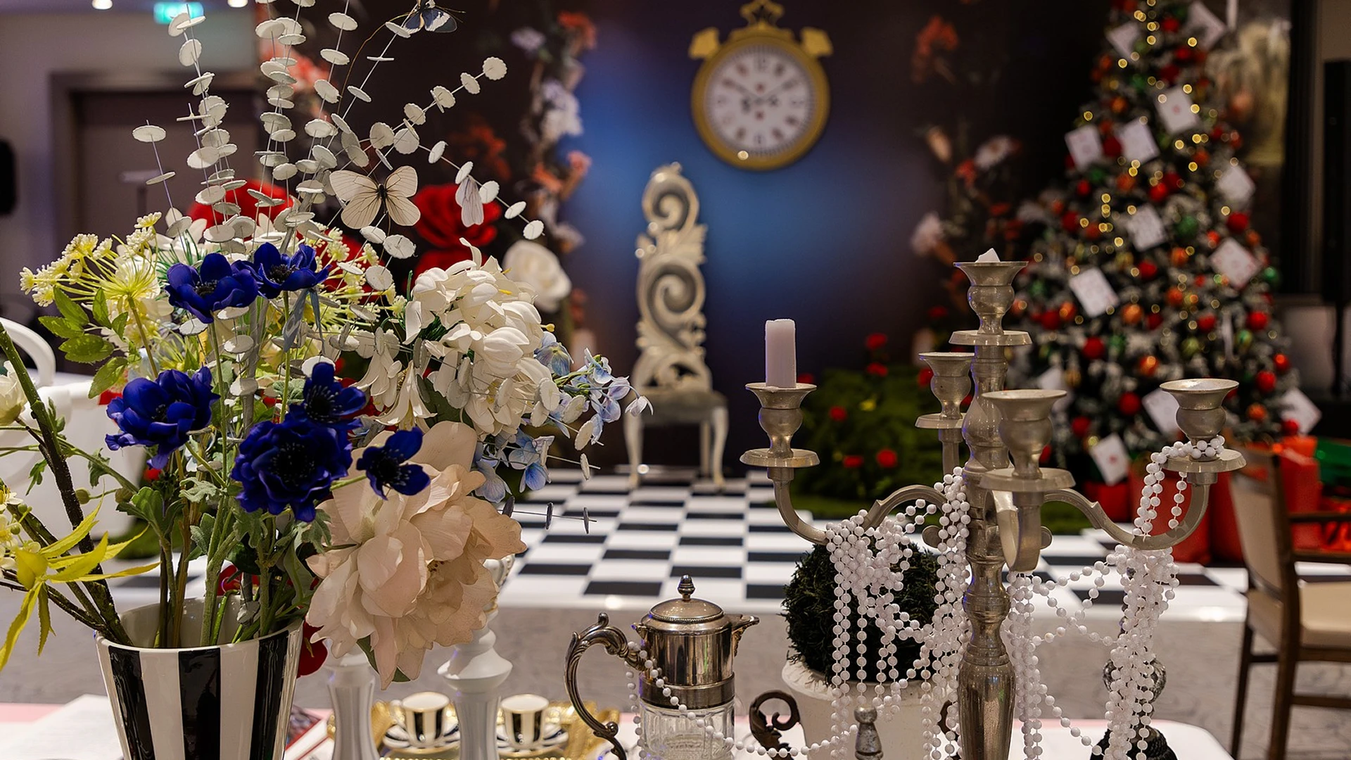 Alice in Wonderland themed corporate event decor featuring a black and white checkered floor, an oversized clock, a candelabra with pearls, and a Christmas tree in the background.