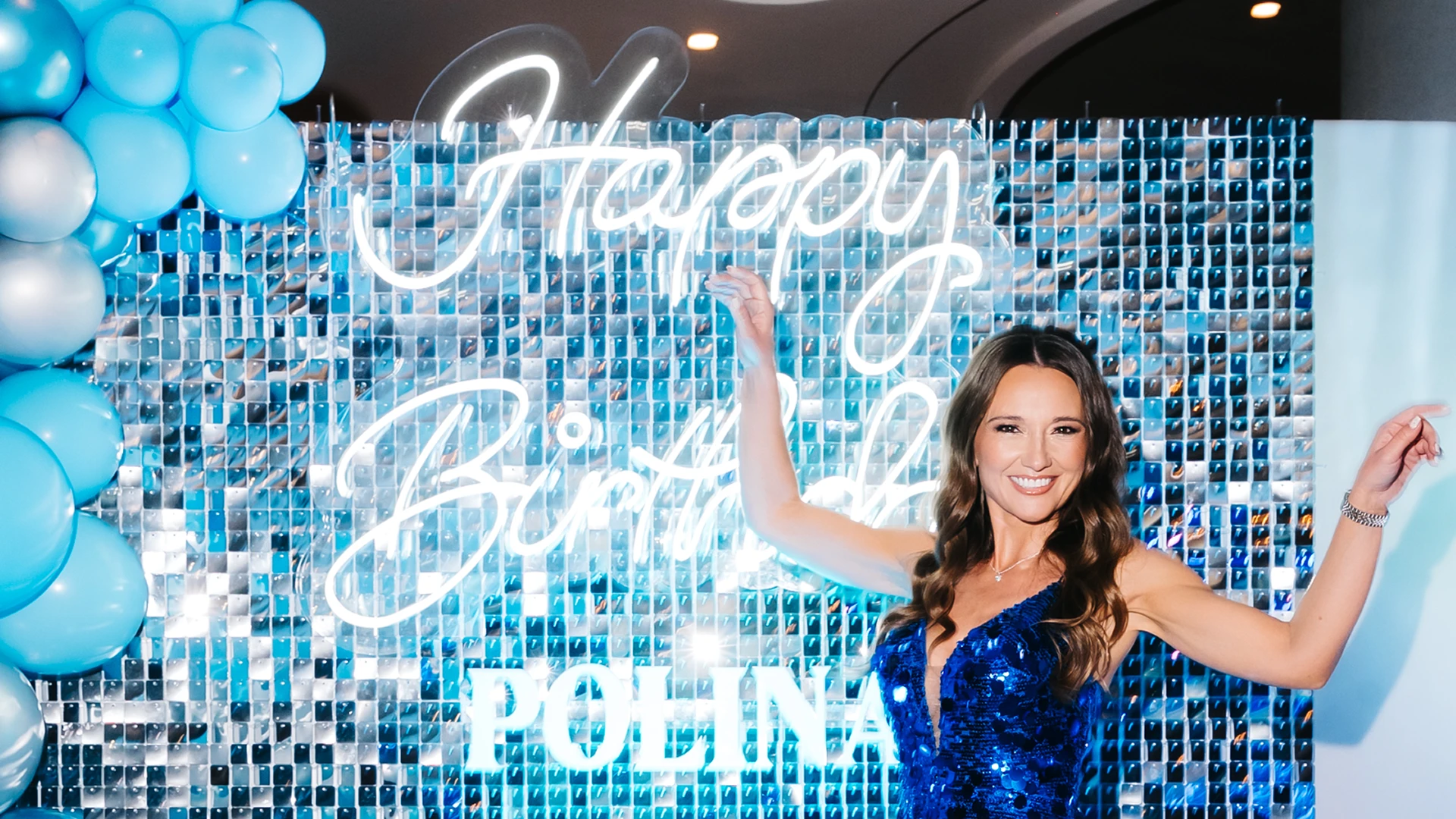 A happy woman in a blue sparkly dress posing in front of a shimmering blue sequin wall with balloons and a personalized neon Happy Birthday sign.