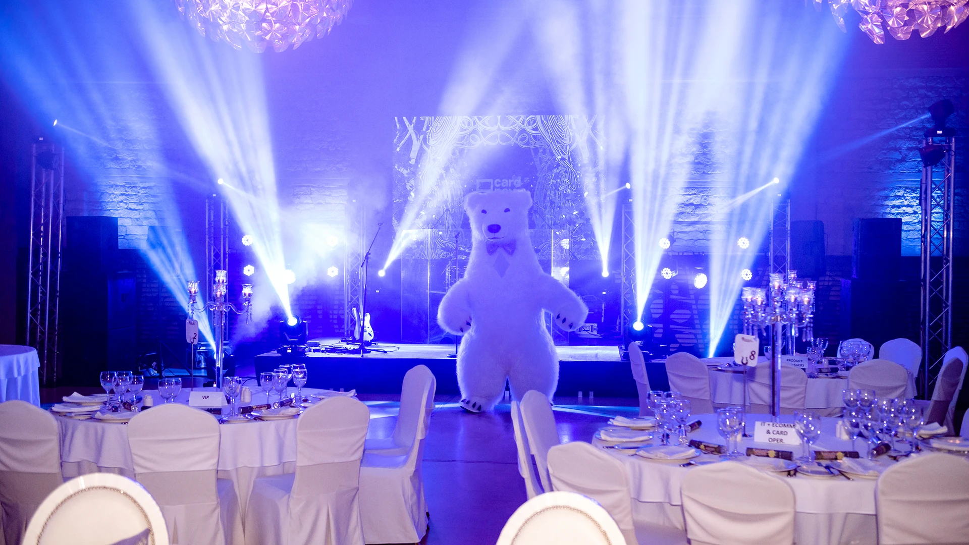 The Great White Bear mascot standing on a stage illuminated by blue spotlights during a formal corporate banquet dinner.