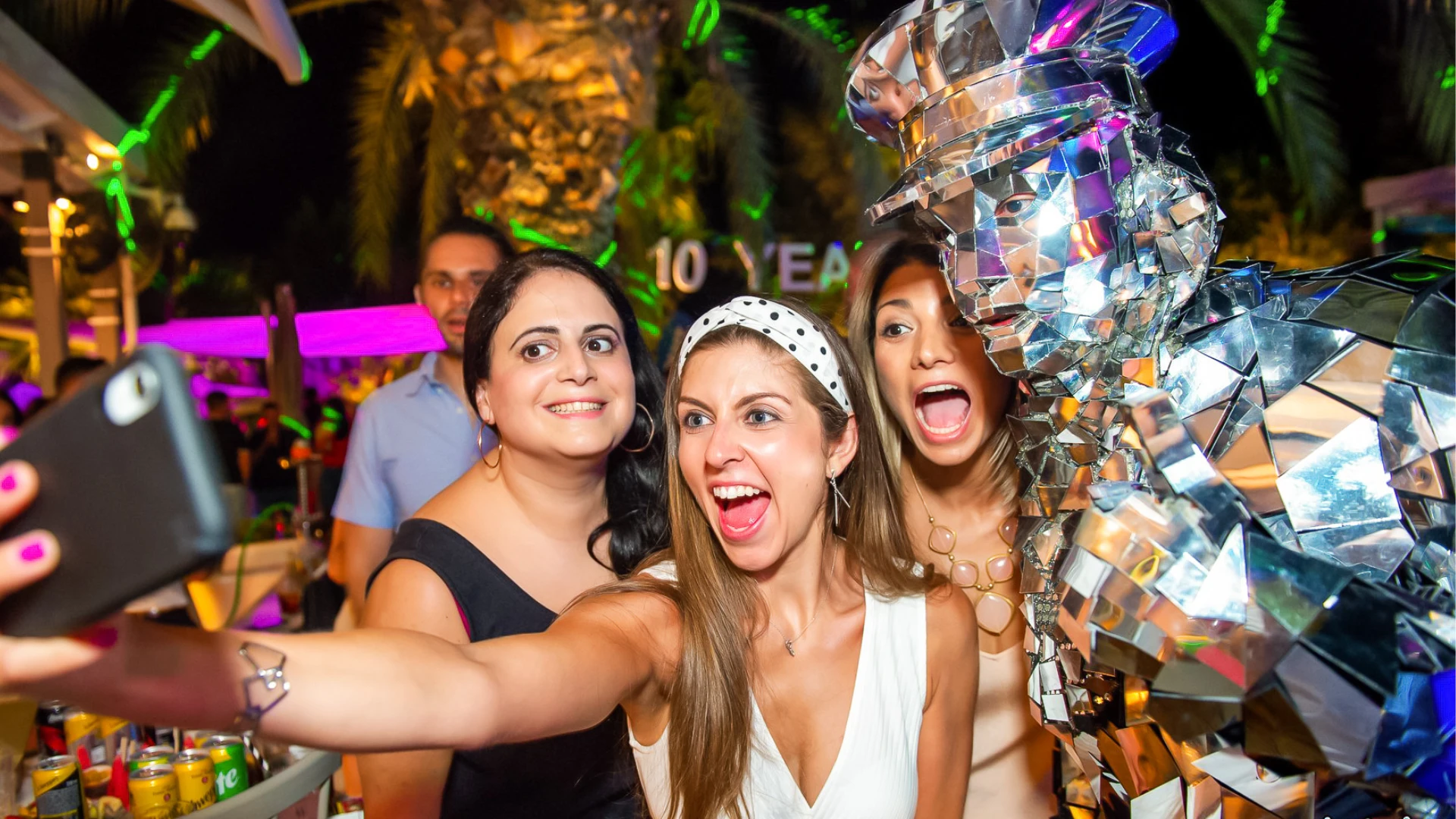 Three excited female guests taking a fun selfie with a sparkling Mirror Man performer during an outdoor evening private party.