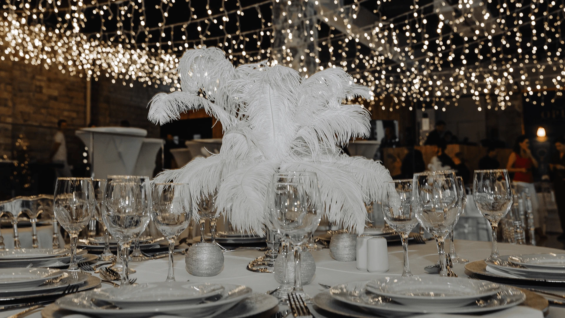 Round table set with glassware and a tall white ostrich feather centrepiece under a fairy-light canopy
