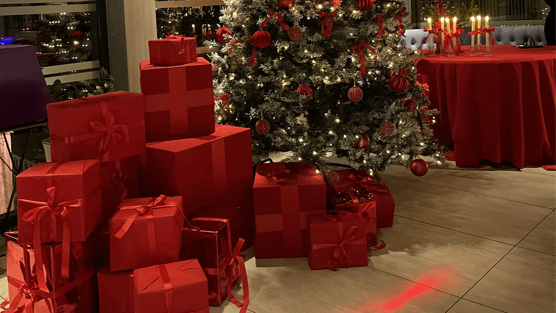 Stack of red wrapped gift boxes beside a decorated tree at an evening corporate event