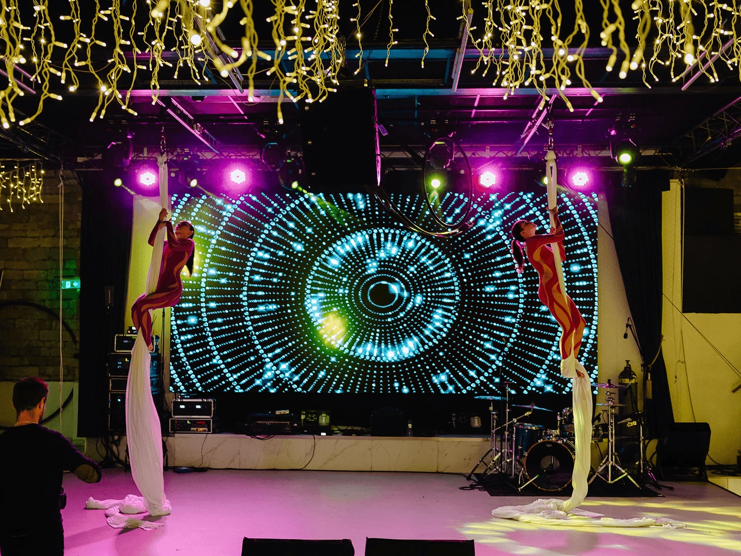 Two aerial silk performers mid-routine on either side of a large LED screen in a decorated hall