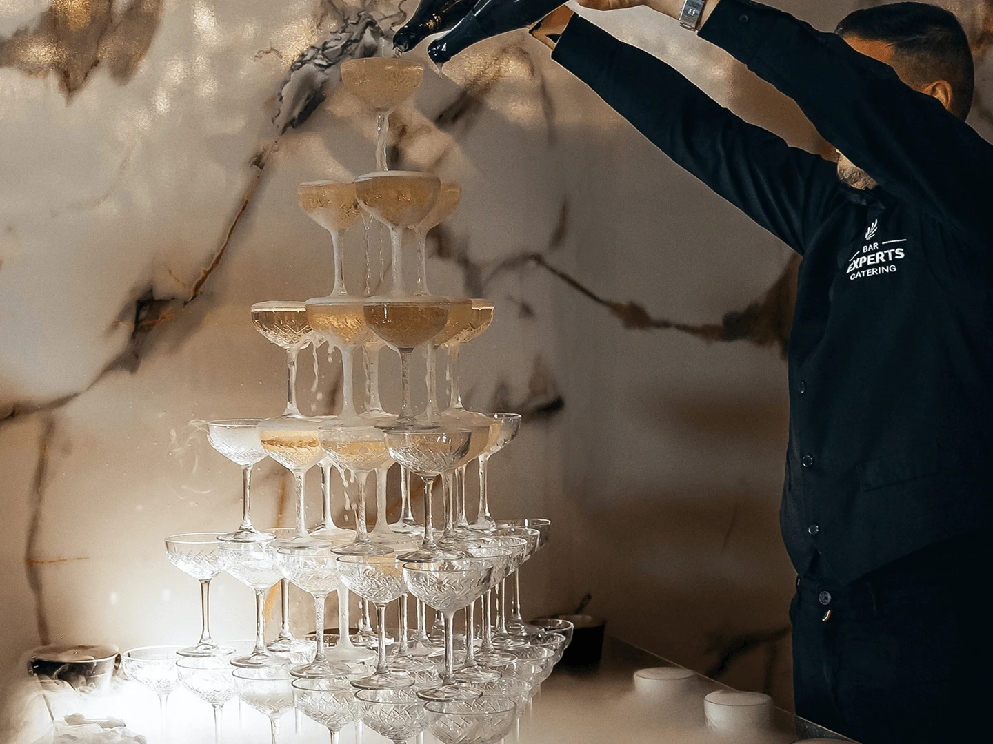 Bartender pouring a champagne tower built from coupe glasses against a marble wall