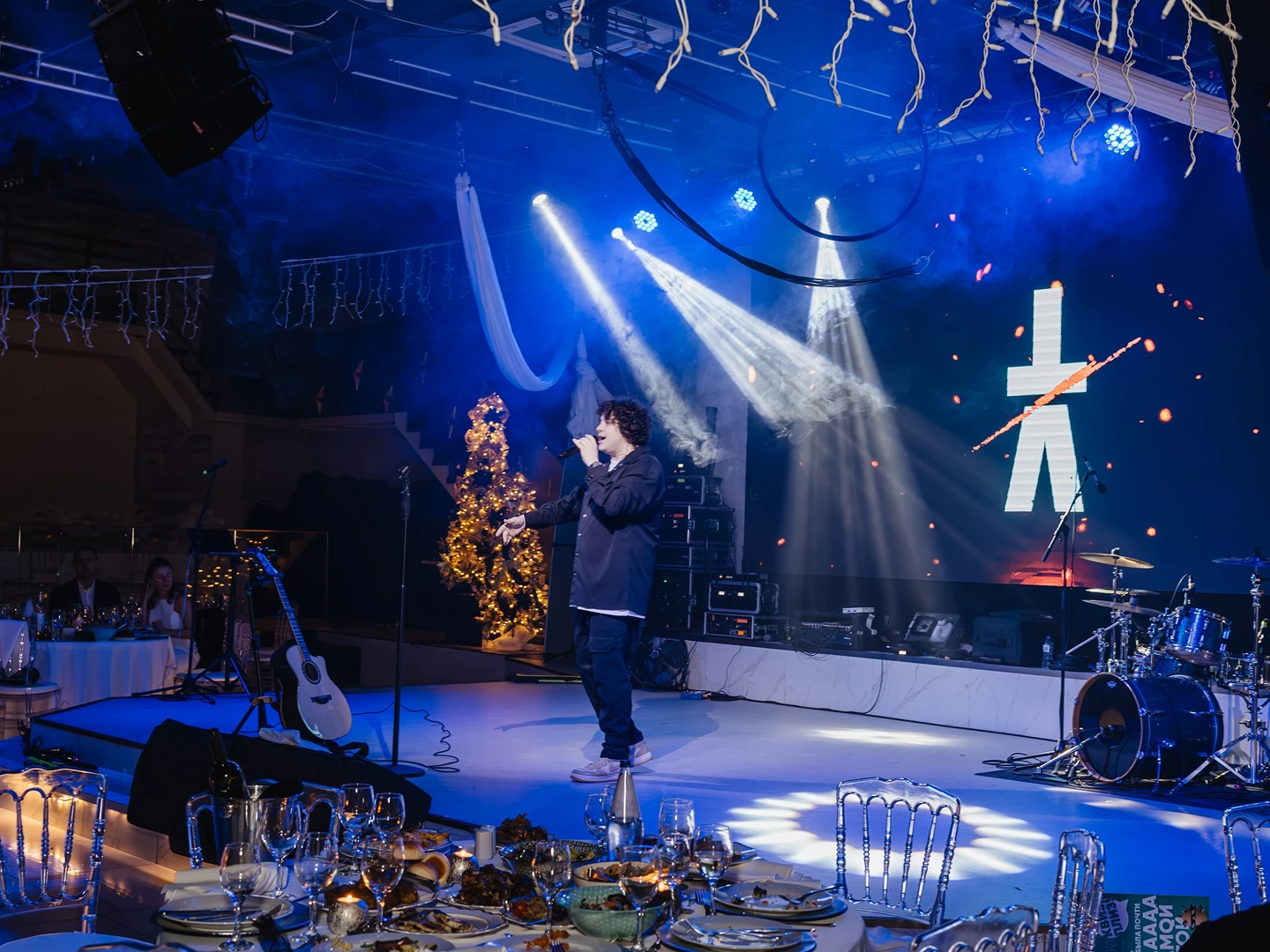 Vocalist singing on a lit stage with a band setup and decorated tree while guests watch from banquet tables