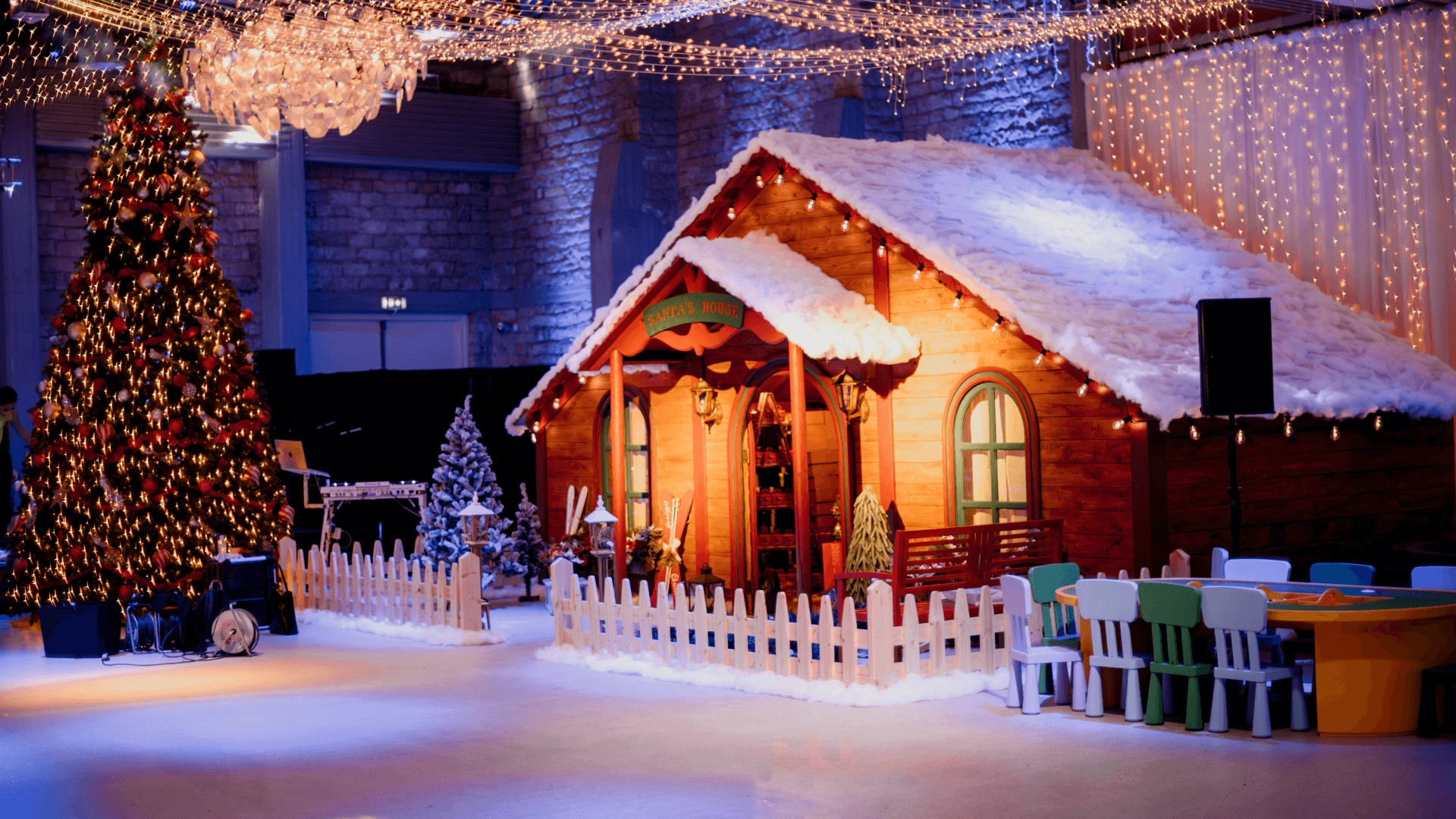 Wooden winter cabin installation with a snow-covered roof, decorated tree and fairy-light canopy indoors