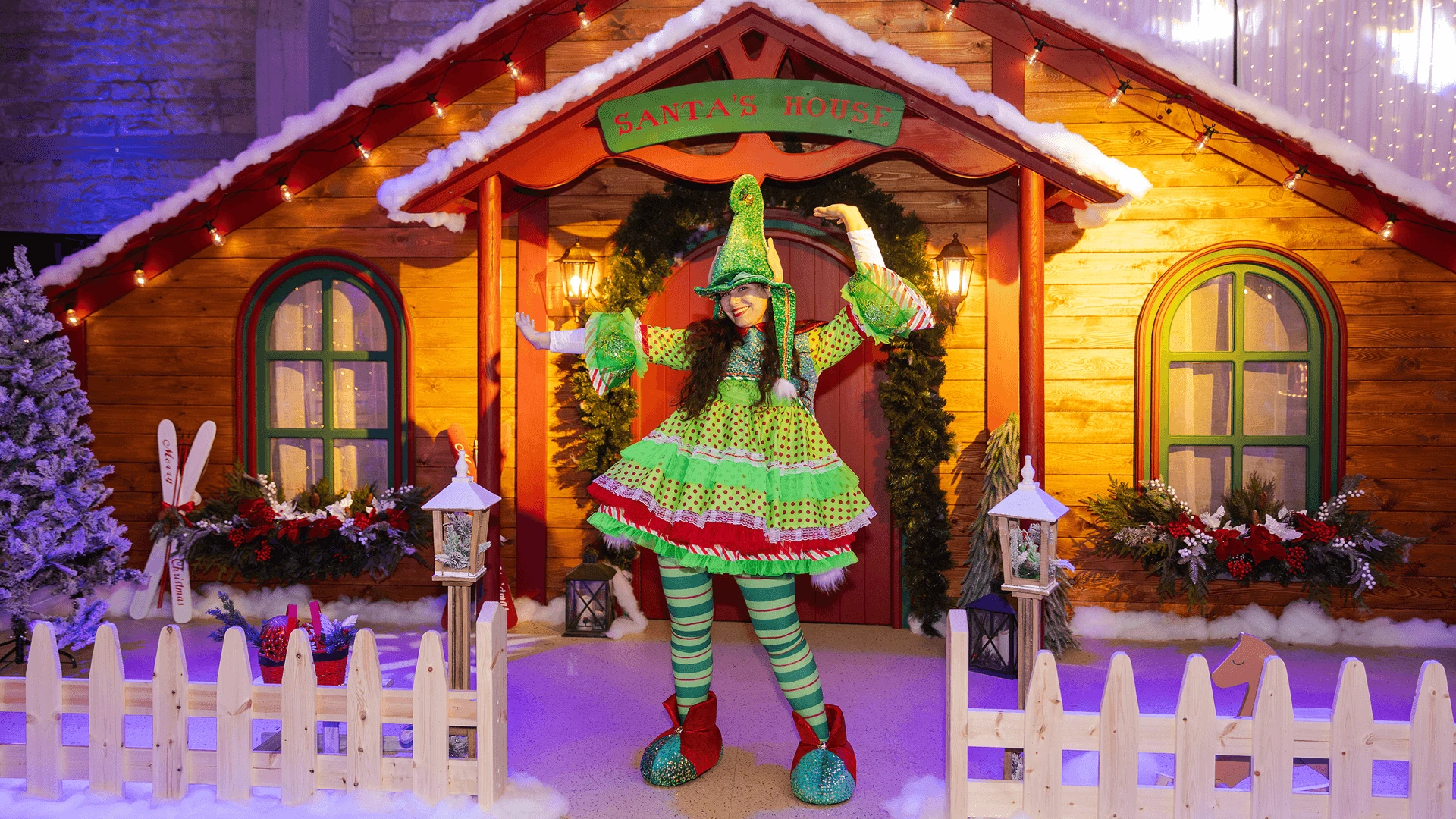 Elf performer posing in front of a decorated wooden cabin set with lanterns and a picket fence