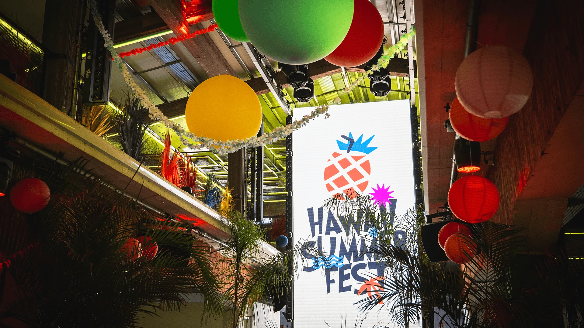 Branded event screen surrounded by oversized balloons, paper lanterns and palm foliage at a themed corporate party