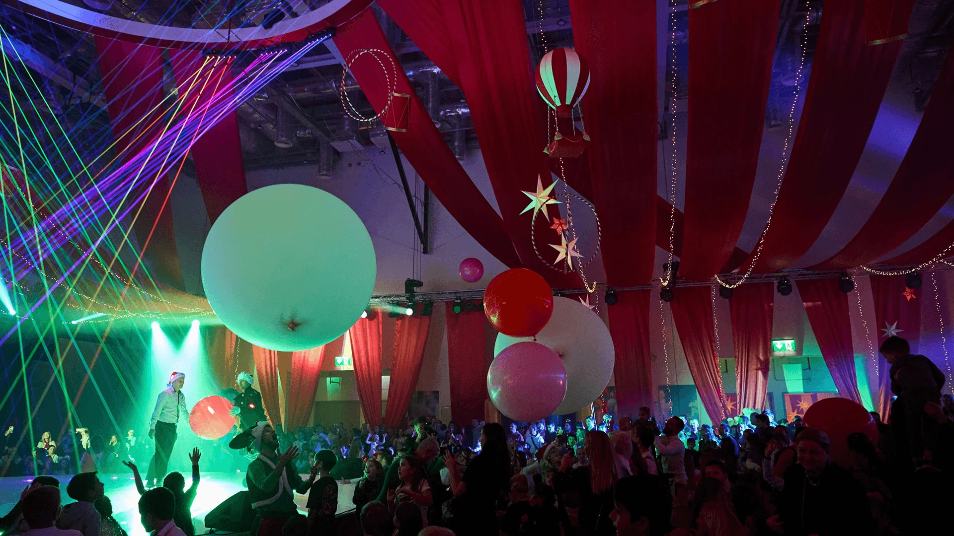 Laser beams and oversized balloons above a large crowd in a hall draped in red and white fabric