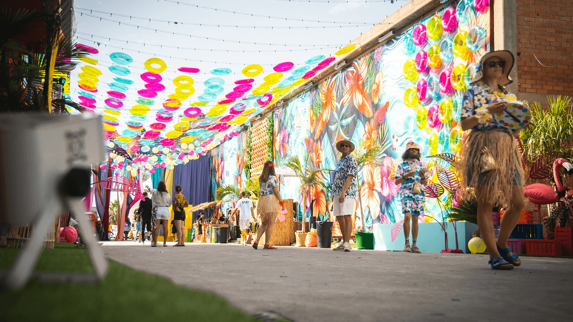 Guests in tropical costume walking through an outdoor venue decorated with a canopy of inflatable rings and a painted mural